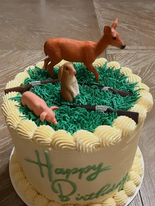 Hunting themed birthday cake topped with a deer, rabbit and rifles in green buttercream grass